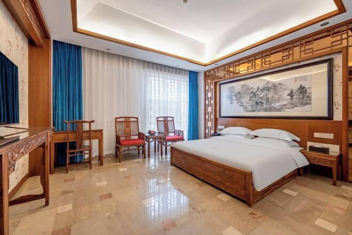 A hotel room with a wooden bed, a desk, two chairs, and a large framed artwork on the wall.