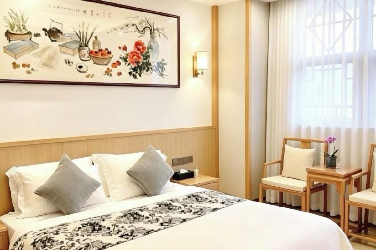 Habitación de hotel con cama, mesitas de noche, una silla, una pequeña mesa con un florero y una ventana con cortinas.