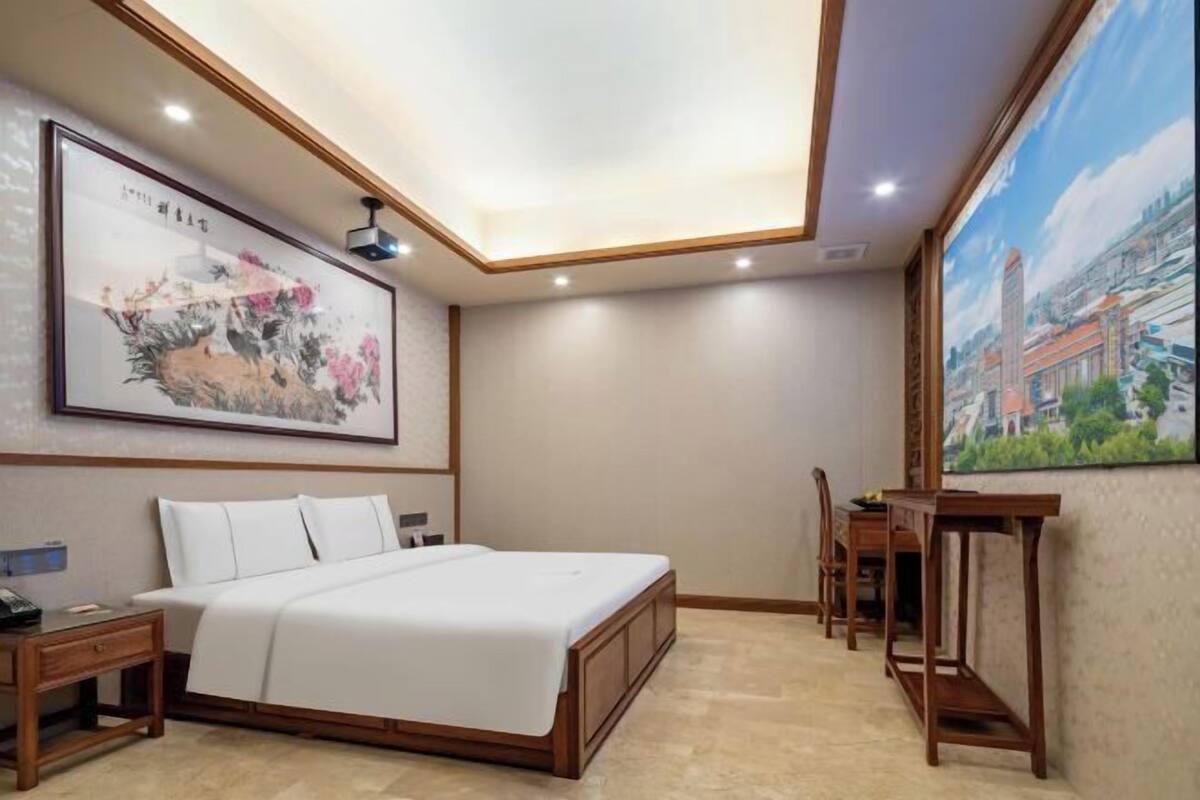 A hotel room with a bed, a desk, a chair, a painting on the wall, and a ceiling light.