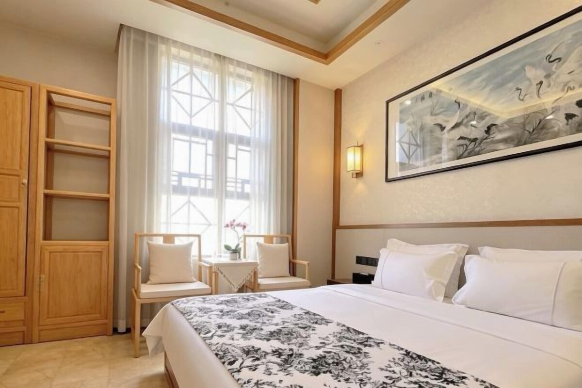Ein Hotelzimmer mit einem großen Bett, einem Schreibtisch mit Stühlen, einem Fenster mit Vorhängen und einem Gemälde an der Wand.