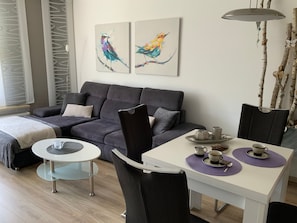 Un salon moderne avec un canapé gris, une table basse en verre, une table à manger blanche avec des chaises, et deux tableaux représentant des oiseaux, accrochés au mur.