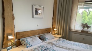 Een opgemaakt bed met beddengoed met een patroon, een houten hoofdbord, nachtkastjes met lampen, een ingelijste foto aan de muur en een raam met gordijnen.