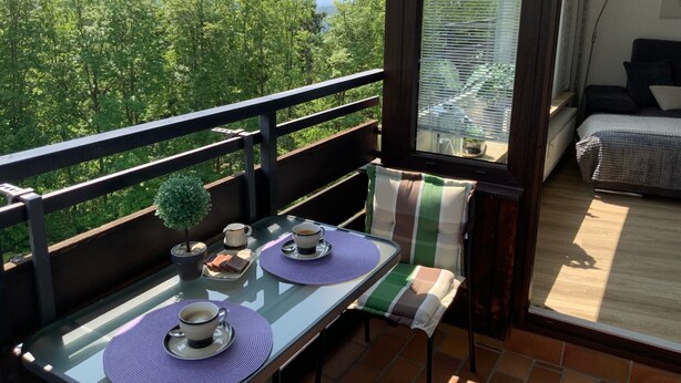 Een balkon met een tafel gedekt voor twee personen, een kleine bloempot en uitzicht op een bebost gebied.