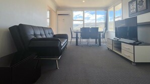 A modern living room with a black leather sofa, a TV on a white cabinet, and a dining area with chairs. - The Mountain Riveting House at Risdon vale (Risdon Vale)