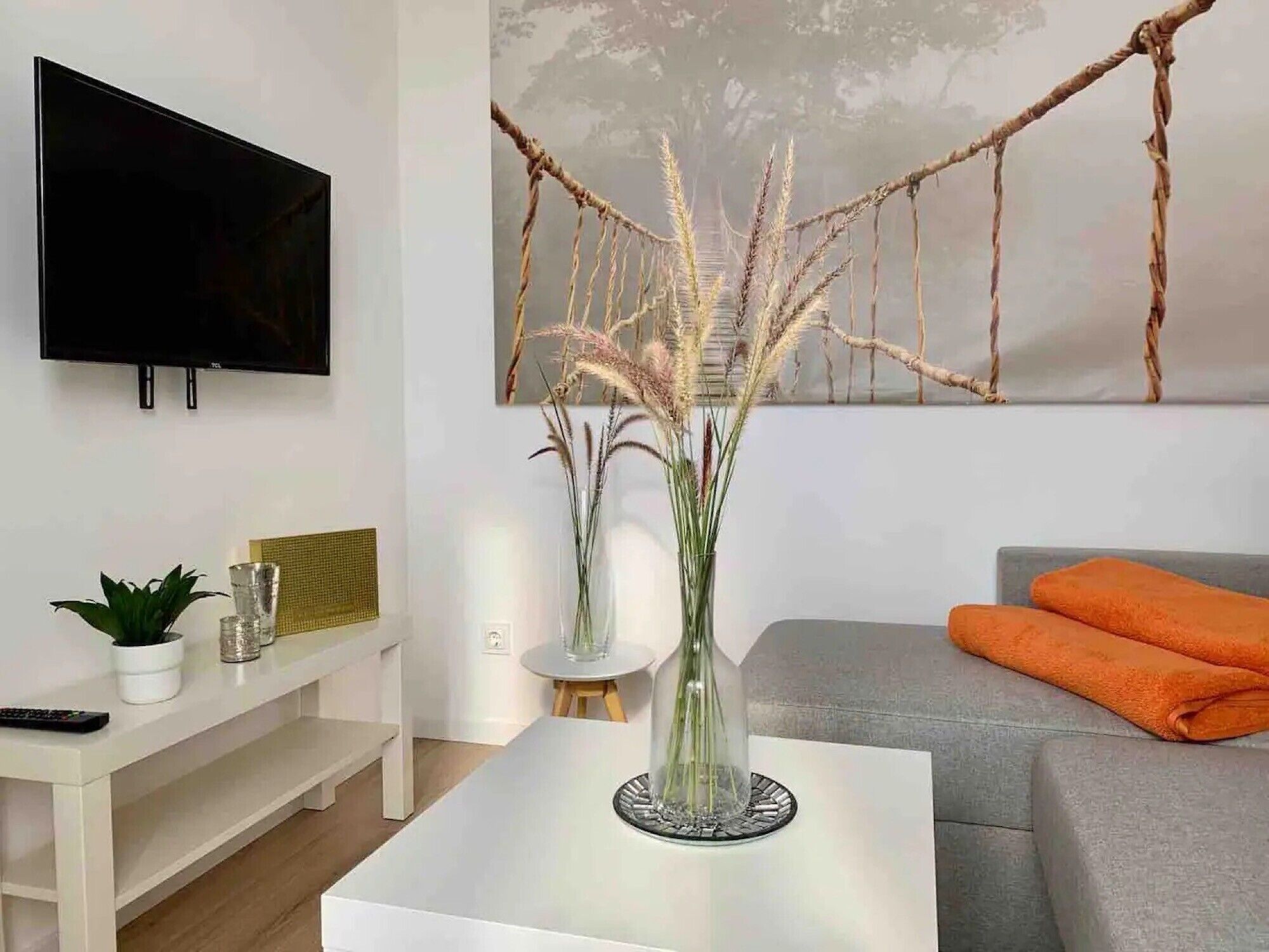 A living room with a flat-screen TV, a white coffee table with a vase, a grey sofa, and a painting on the wall.