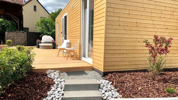Ein Holzhaus mit Terrasse, Sitzgelegenheit im Freien und einem kleinen Garten.