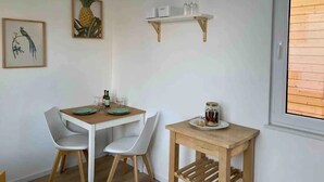 Ein Essbereich mit einem weißen Tisch und Stühlen, einem kleinen Holznachttisch mit einer Glaskaraffe und einem Fenster mit Holzjalousien.