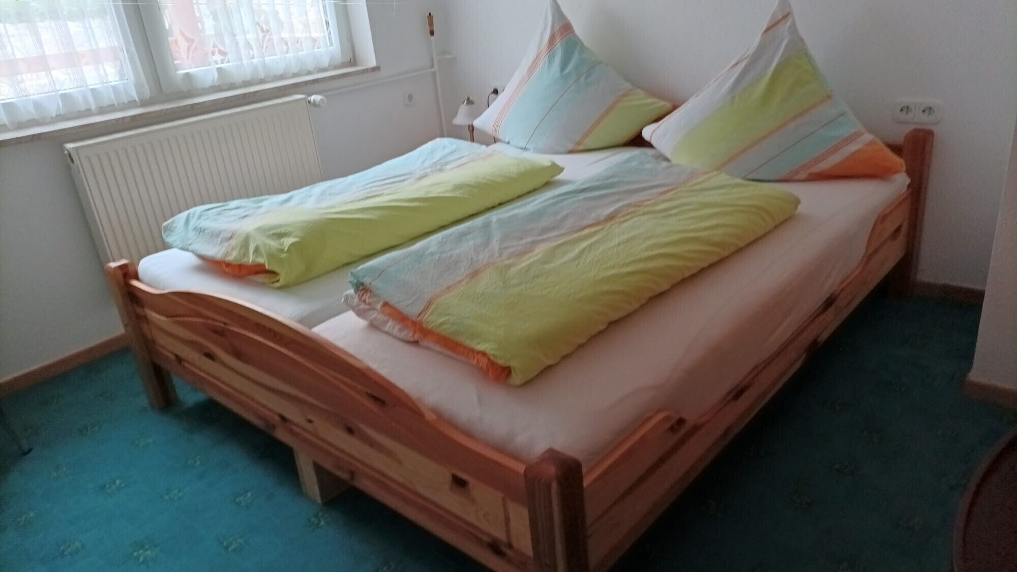 Uma cama de solteiro com estrutura de madeira, um radiador e uma janela com persianas.
