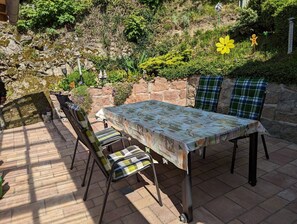 Une terrasse extérieure avec une table et des chaises, entourée d’un mur de pierre et de verdure.