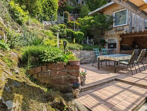 Une terrasse avec une table et des chaises, entourée d’une végétation luxuriante et d’un mur de pierre.