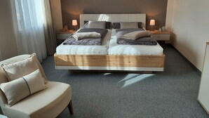 A double bed with a wooden frame, two bedside tables with lamps, a chair, and a large window with sheer curtains. - Apartment "Am Kapellenberg" with Mountain View, Shared Terrace & Wi-Fi (Bad Peterstal-Griesbach)