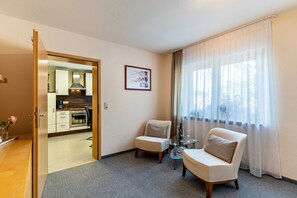 A hotel room with a kitchenette, a dining area with a table and two chairs, a large window with sheer curtains, and a framed picture on the wall.