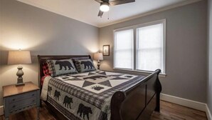 Een slaapkamer met een bed, nachtkastje, lamp, plafondventilator en raam met jaloezieën.