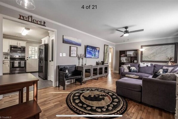 A modern living room with a grey sofa, a coffee table, and a patterned rug. There is a kitchen visible in the background with white cabinets and appliances.