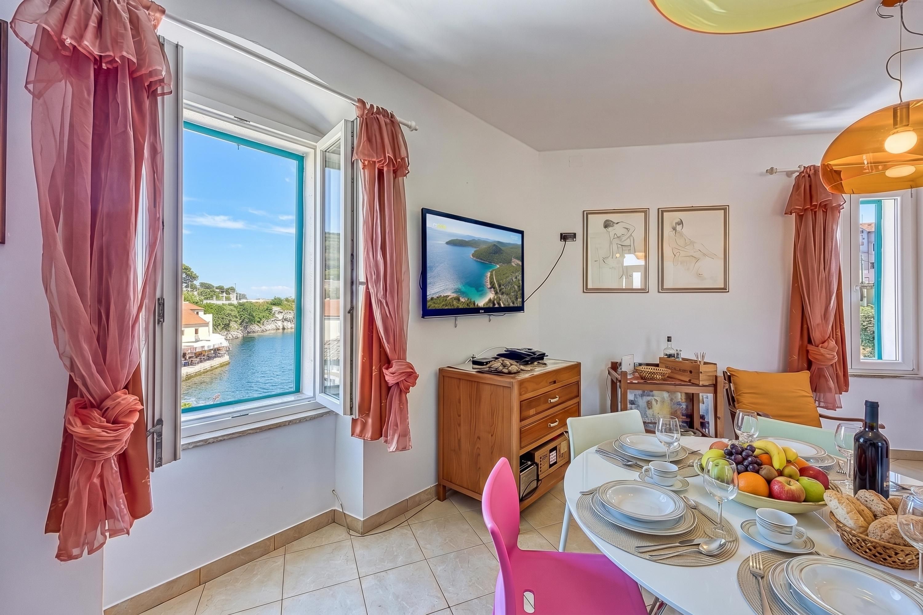 Un coin repas avec une table dressée pour deux personnes, un téléviseur fixé au mur et une vue sur un plan d’eau par une fenêtre ouverte.