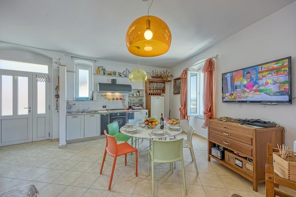 Cozinha moderna com mesa de jantar, cadeiras coloridas, televisão e armário de madeira.