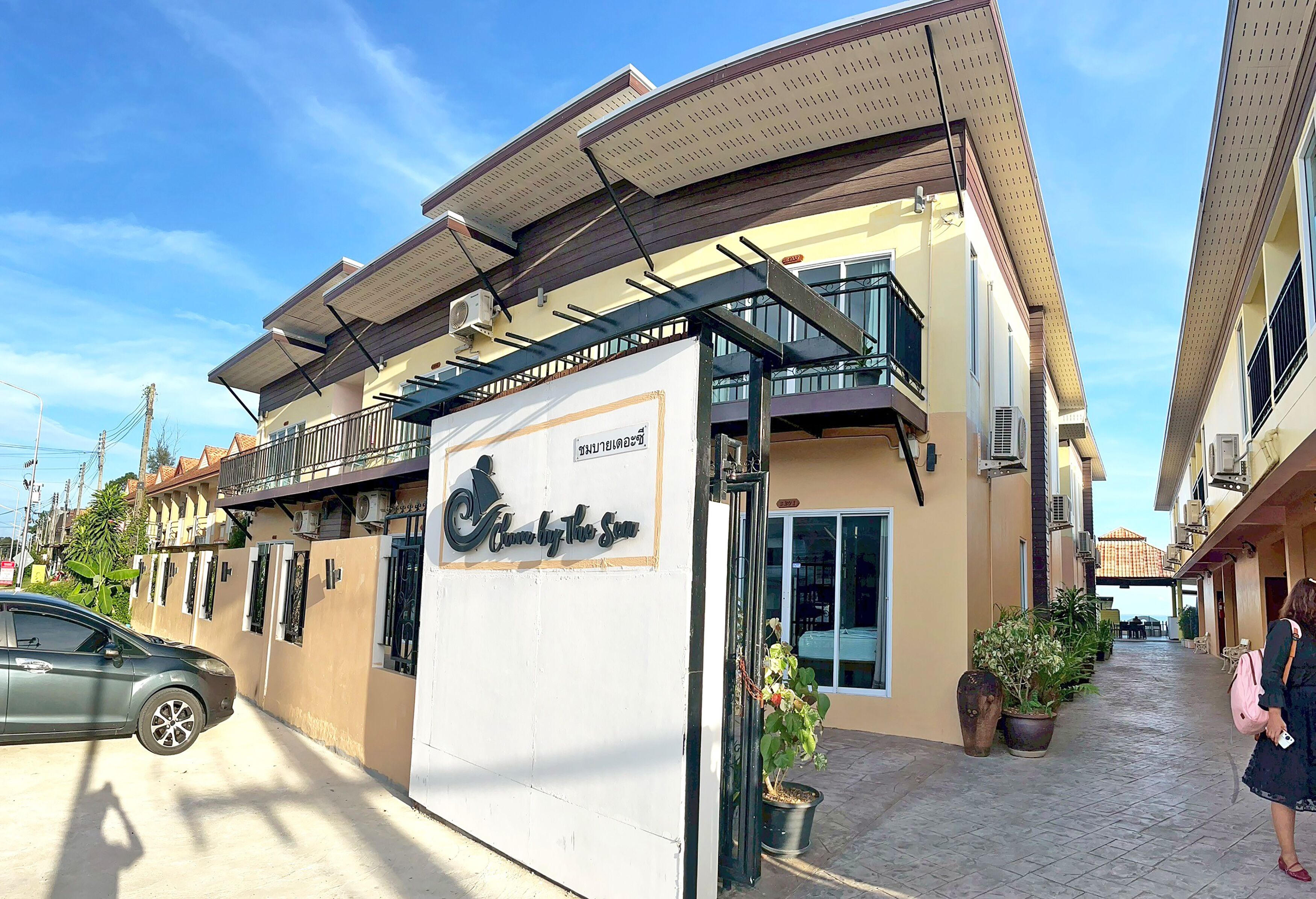 A hotel with a sign reading "Hotel by the Sea." It features a balcony, potted plants, and a clear sky.
