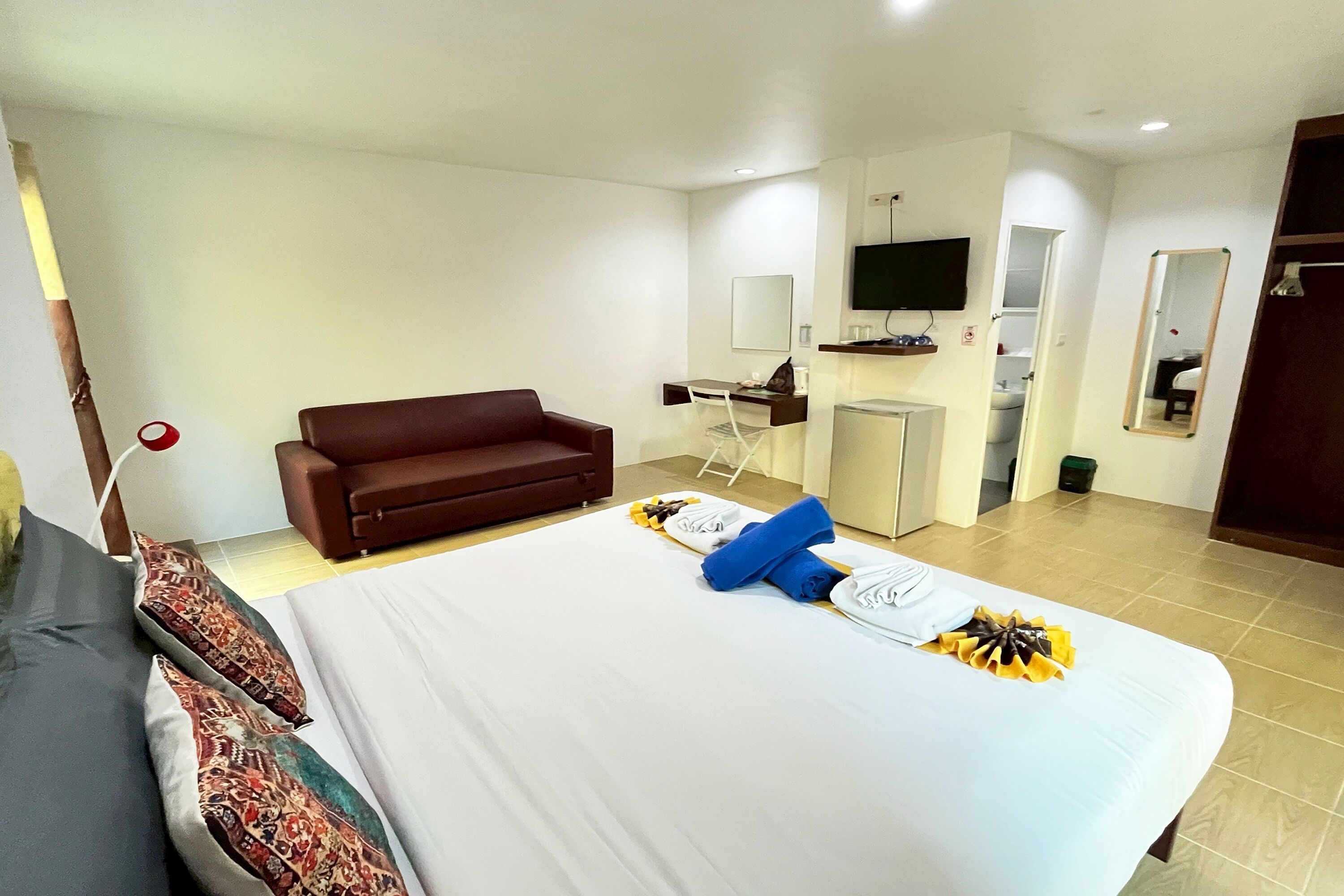 Una camera d'albergo con un letto, un divano marrone, una scrivania con una sedia, una televisione e un bagno visibile attraverso una porta aperta.