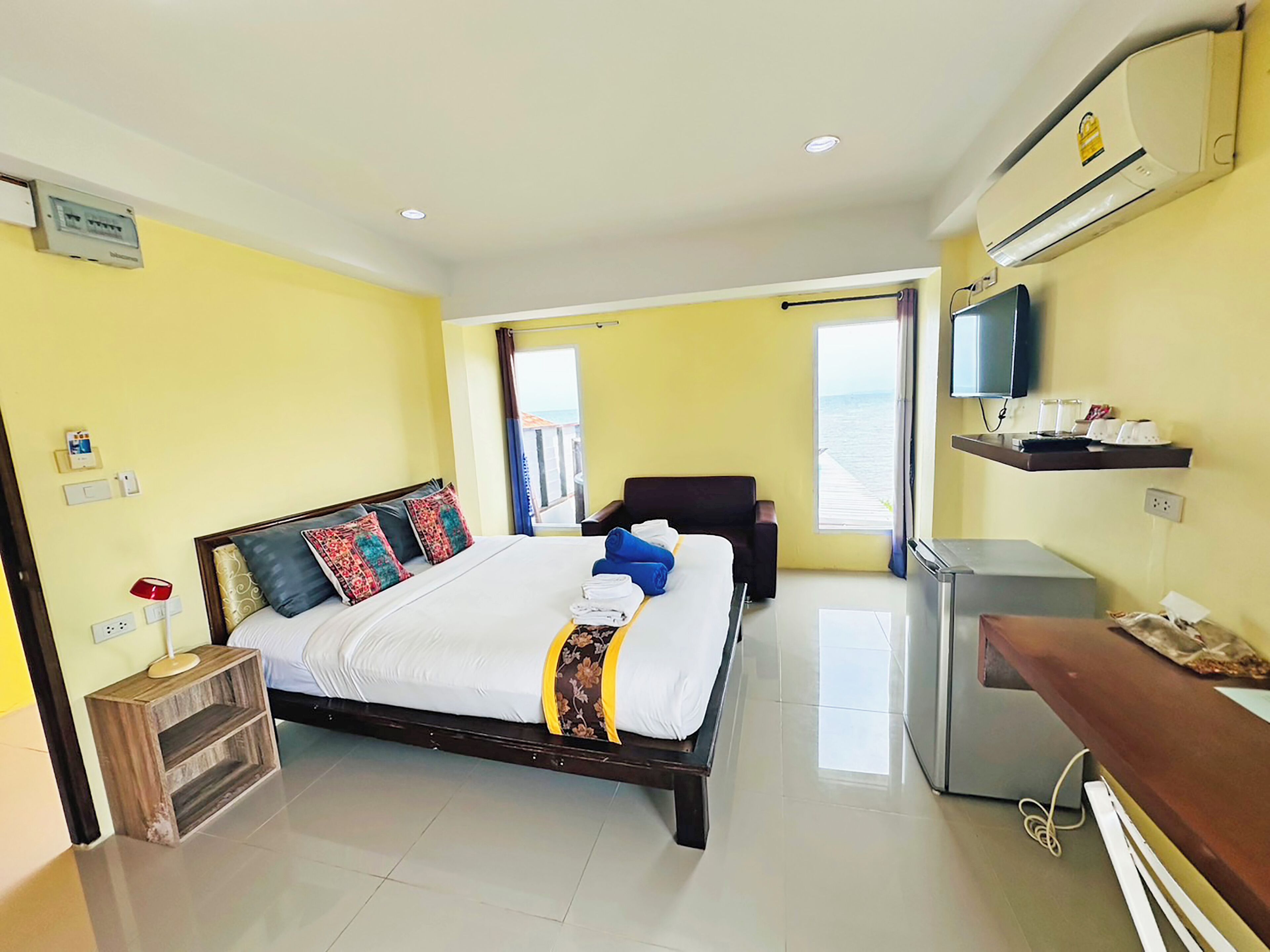 A hotel room with a bed, bedside table, TV, air conditioning unit, and a view of the sea.