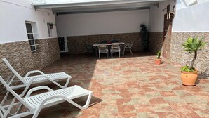 Un patio techado con suelo de baldosas, sillones blancos, una mesa con sillas y plantas en macetas.