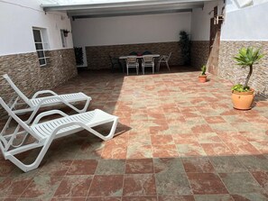 A covered patio area with a tiled floor, white lounge chairs, a table with chairs, and potted plants.