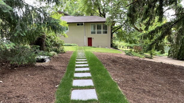 Ein Weg führt zu einem Haus mit roter Tür, umgeben von gepflegtem Rasen und Bäumen.