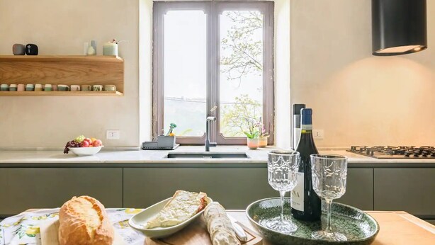 Une cuisine avec une fenêtre, un évier, une cuisinière et une table sur laquelle se trouvent une miche de pain, une assiette de nourriture et deux verres à vin.