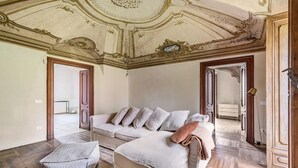 Une pièce dotée d’un grand plafond richement décoré, d’un canapé beige agrémenté de nombreux coussins, d’un pouf à motifs et d’un parquet en bois.
