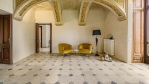 Une pièce avec deux fauteuils jaunes, une petite table ronde, un lampadaire et un radiateur. Le sol est recouvert de carreaux formant un motif géométrique, et le plafond est orné de moulures travaillées, rehaussées de dorures.