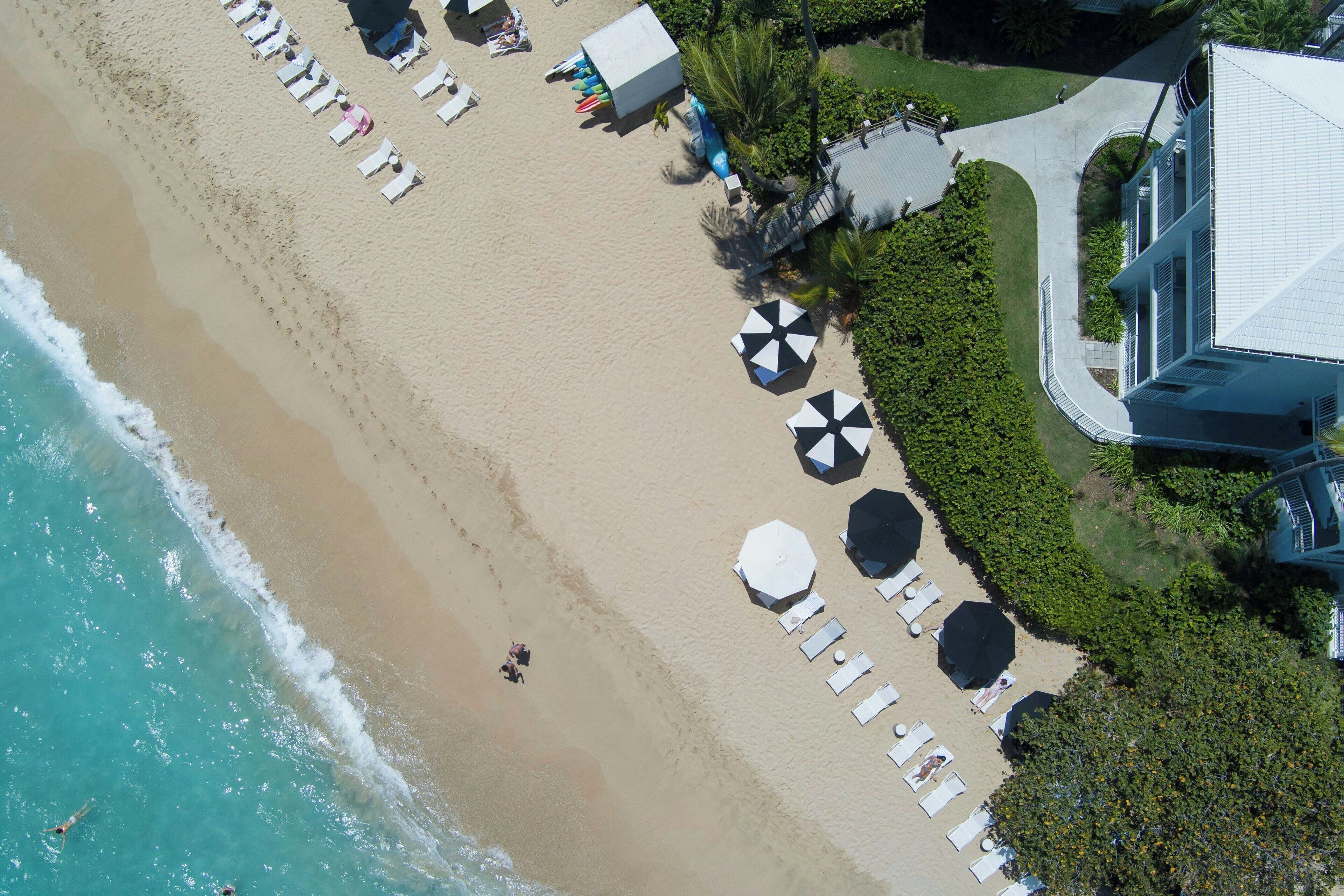 Une vue aérienne d’une plage avec des chaises longues et des parasols, un bâtiment doté d’un toit en pente, et un chemin menant à la plage.
