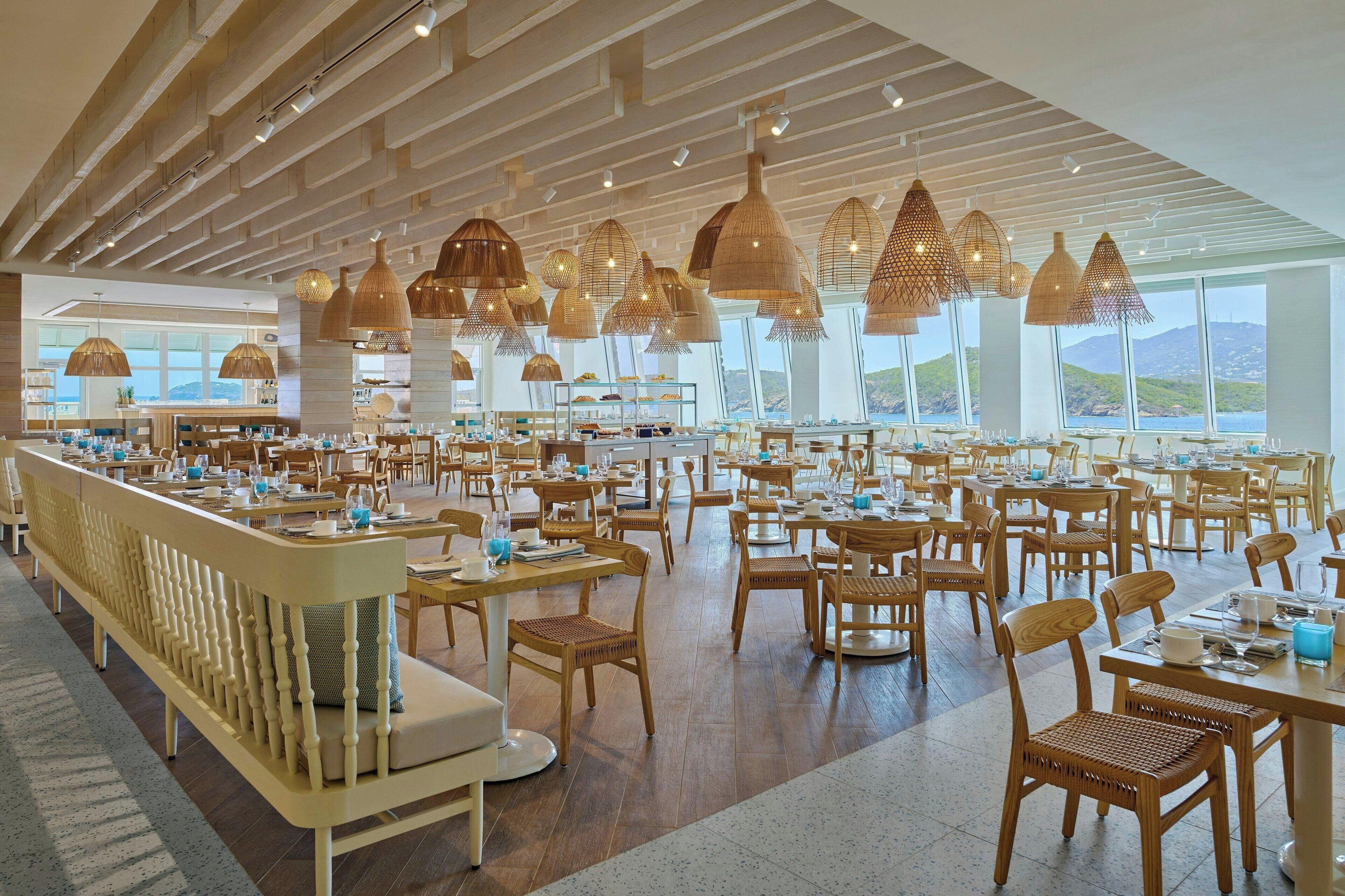 A spacious restaurant with high ceilings, wooden furniture, and large windows offering a view of the hills.