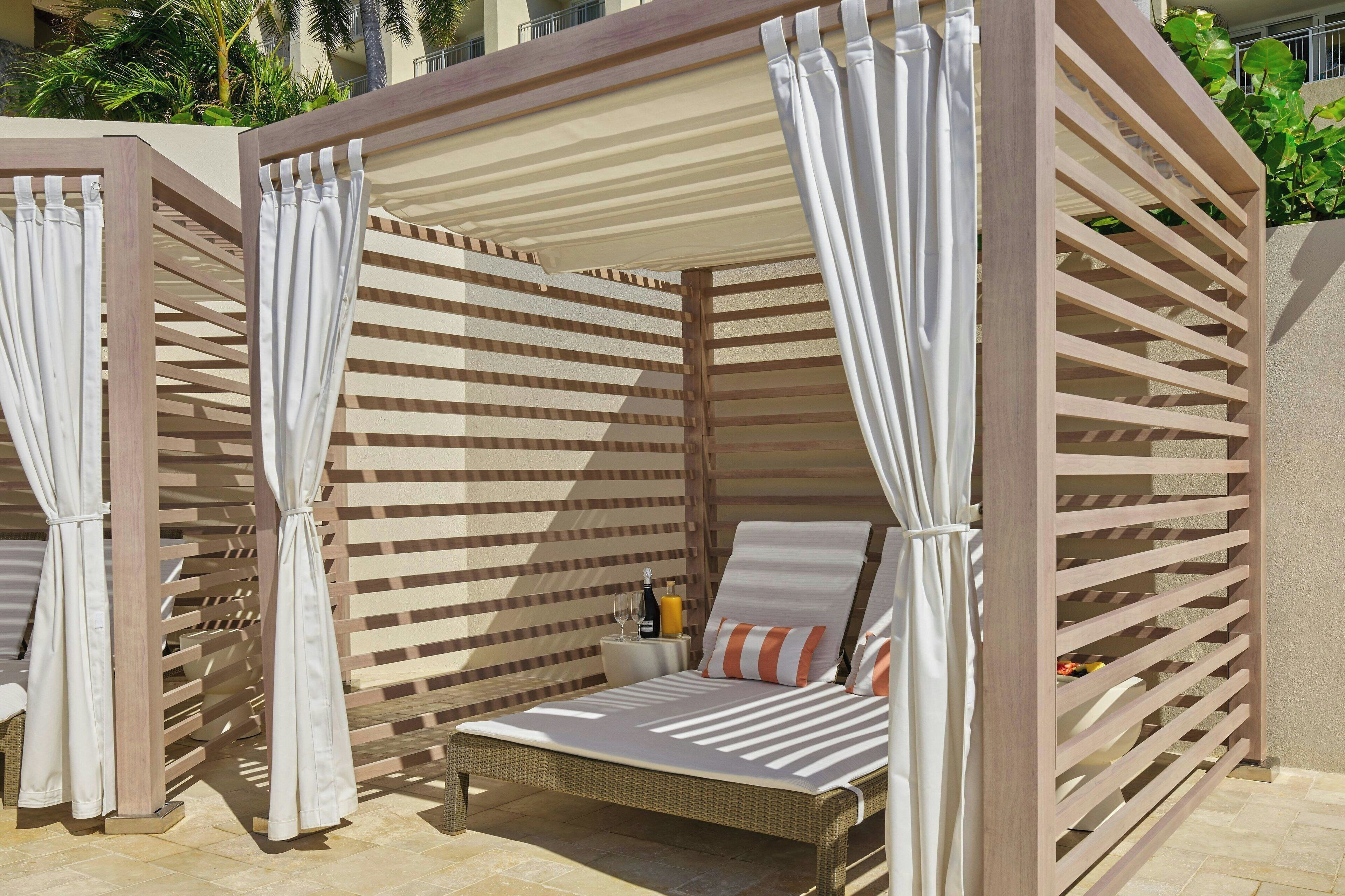 A sunlit outdoor seating area with a wicker lounge chair, a small table with a bottle and glass, and a wooden pergola structure.