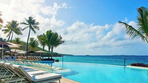 Zona de piscina con sillones y sombrillas, palmeras y vista al mar.