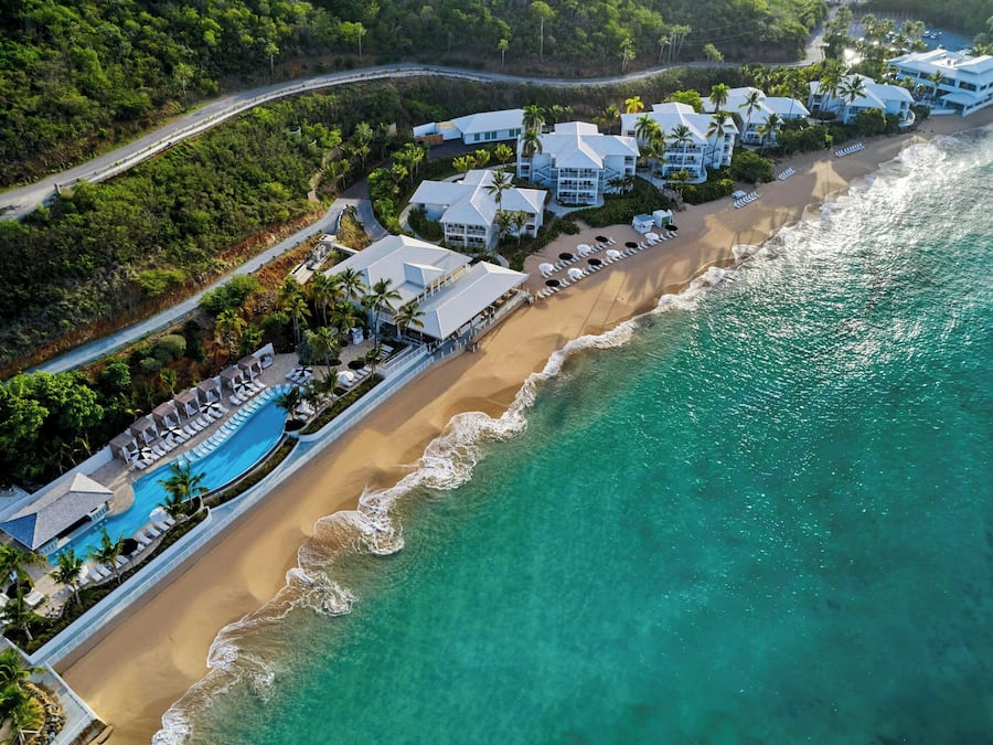 An aerial view of a resort with a long beachfront, a swimming pool, and multiple buildings with white roofs.