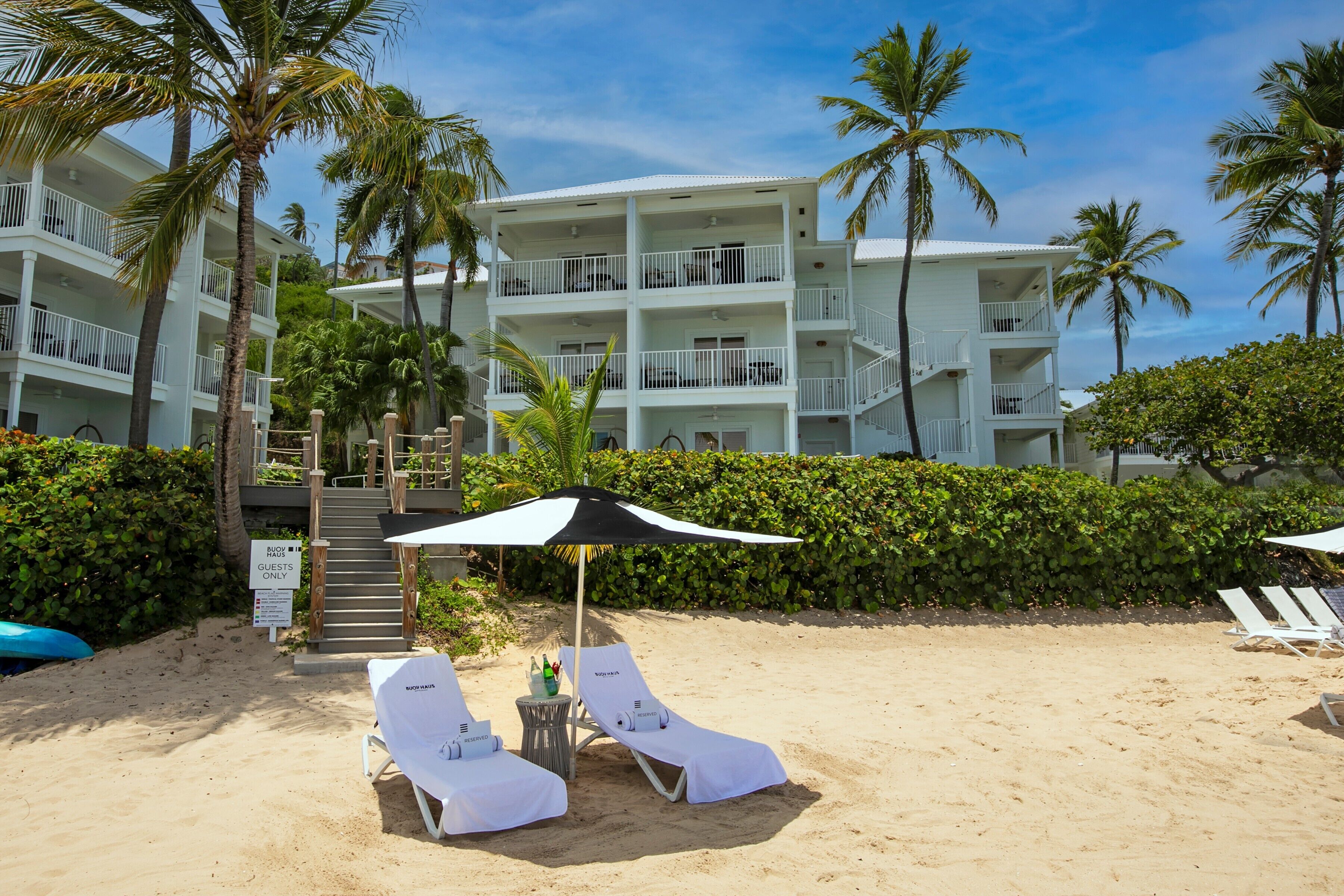 A beachfront hotel with multiple balconies, a sandy beach area with lounge chairs, and palm trees.