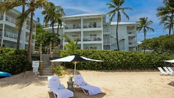 A beachfront hotel with multiple balconies, a sandy beach area with lounge chairs, and palm trees.