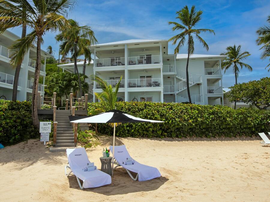 A beachfront hotel with multiple balconies, a sandy beach area with lounge chairs, and palm trees.
