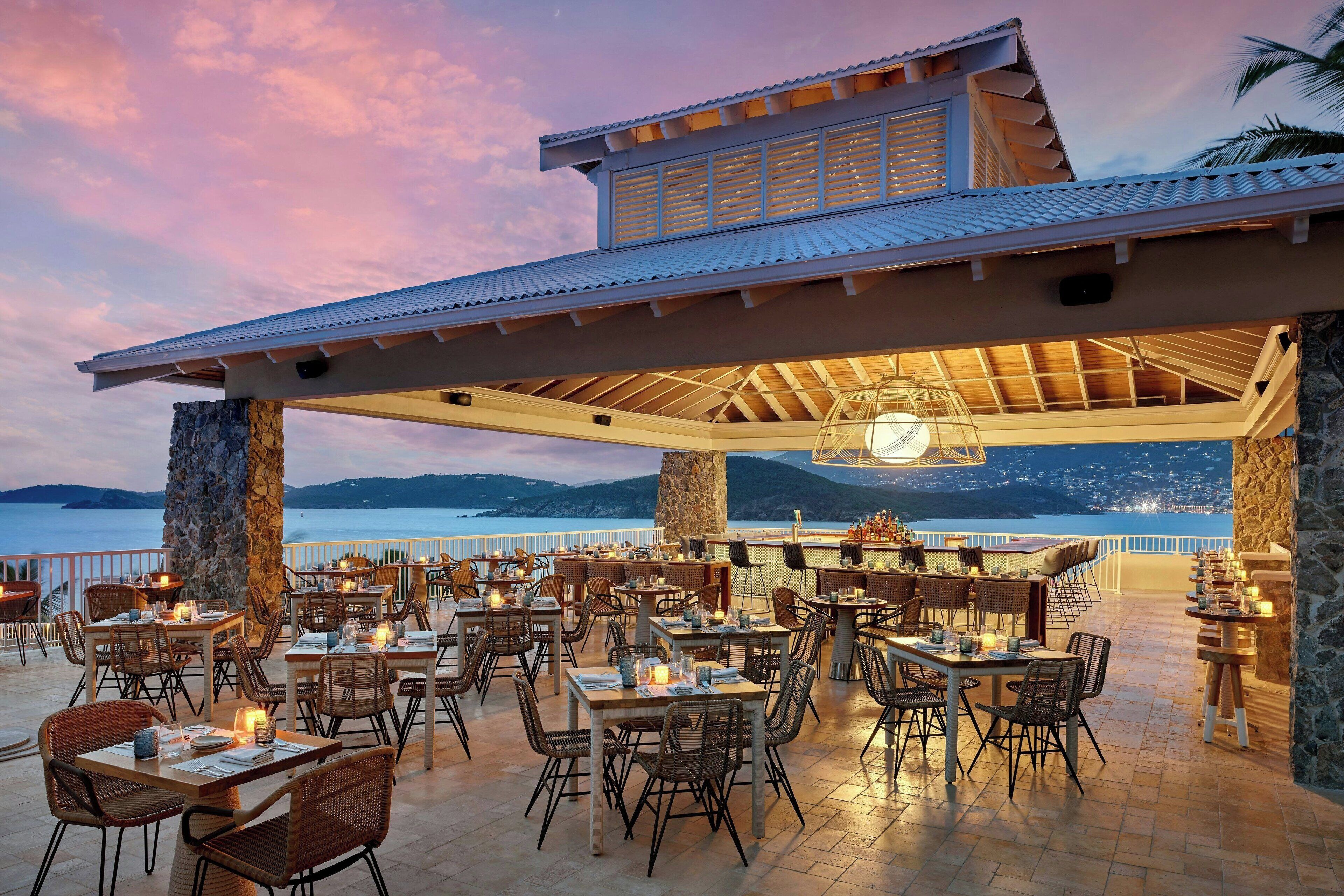 An outdoor dining area with tables and chairs, a stone pillar, and a view of the sea.