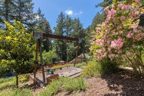 A wooden swing set in a garden with a picnic table and benches, surrounded by trees and shrubs.