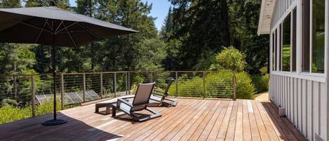 A wooden deck with two lounge chairs and a large umbrella, surrounded by a metal railing and dense trees.