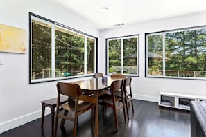 A dining area with a large table and chairs, large windows with views of greenery, and a white wall with a painting.