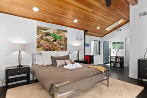 Ein modernes Schlafzimmer mit Holzboden, einem großen Bett mit beiger und brauner Bettdecke, zwei Nachttischen mit Lampen, einem großen Gemälde an der Wand und einem Bücherregal im Hintergrund.
