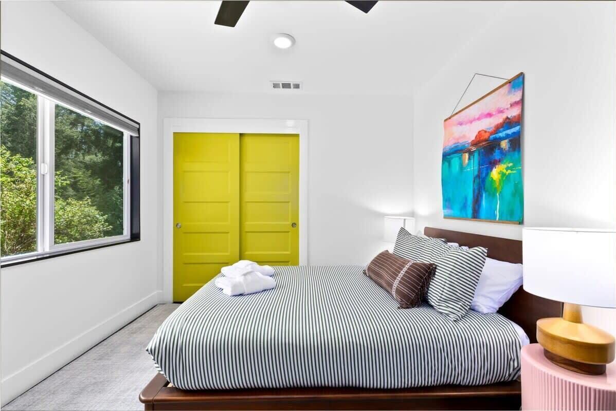A modern bedroom with a large window, a bright yellow door, a bed with striped bedding, and a colorful abstract painting on the wall.