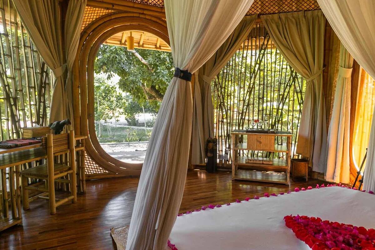 A room with a bed, a table, and chairs, bamboo walls, and a view of greenery.