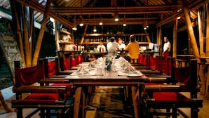 Un restaurant avec des tables en bois et des chaises à coussins rouges, un plafond haut avec des poutres apparentes et une vue sur la cuisine.