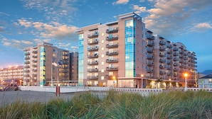 Un hôtel à plusieurs étages, doté de balcons, situé près d’une plage. On aperçoit une clôture et une zone herbeuse au premier plan.