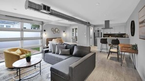 En moderne stue med sofa, sofabord og stole. Der er et køkkenområde med spisebord og stole. Et stort vindue giver udsigt udad.