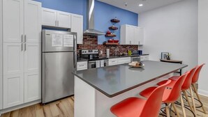 A modern kitchen with white cabinets, a stainless steel refrigerator, a brick backsplash, and a central island with red chairs. - 6x Gamer Themed 2BR Apts - Close to Stadium! (Jacksonville)