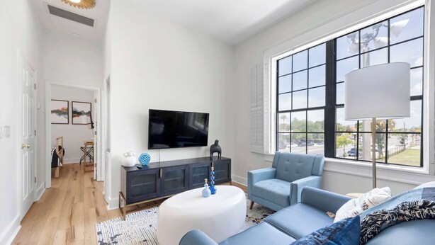 A modern living room with a large window, a flat-screen TV, a sofa, and a coffee table. - 6x Gamer Themed 2BR Apts - Close to Stadium! (Jacksonville)