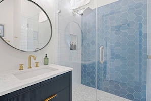 A bathroom with a round mirror, a sink with gold faucets, a glass shower enclosure, and a hexagonal tile pattern on the walls.